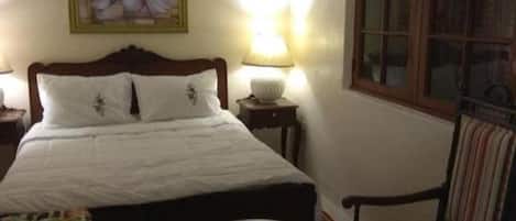 A bedroom with a bed, a chair, a small table, a lamp, a painting, and a window.
