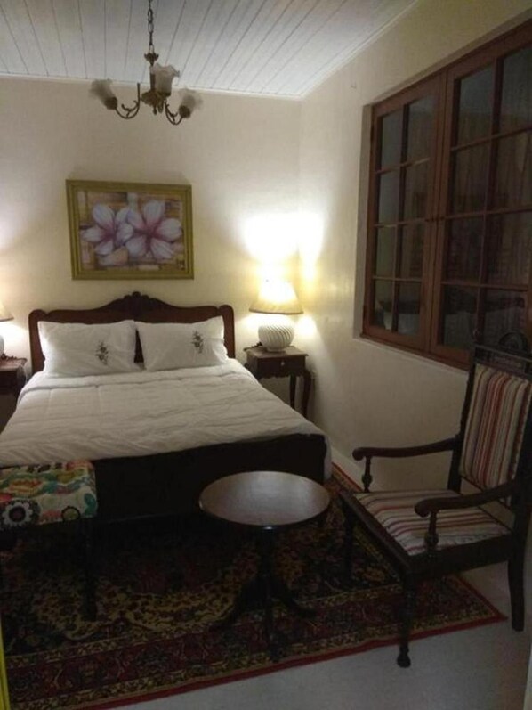 A bedroom with a bed, a chair, a small table, a lamp, a painting, and a window.