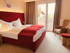 Una habitación de hotel con una cama grande, un cabecero rojo, una mesita de noche de madera, un sillón rojo y vistas al exterior a través de una puerta corrediza de vidrio.