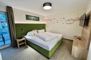 Habitación de hotel con cabecera verde, cama con ropa blanca, mesita de noche, televisor montado en la pared y ventana con cortinas.