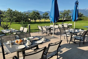 Un espacio de comedor al aire libre con mesas y sillas, sombrillas azules y vistas a montañas.