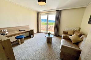 Quarto de hotel com escrivaninha de madeira, cadeira azul, sofá marrom, mesa redonda pequena e vista para o exterior através de uma porta de correr de vidro.