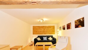 A room with a wooden ceiling, a black sofa with cushions, a white chair, a wooden table, and a wall-mounted light fixture. - Ferienhaus In Bayern Mit Waldblick (Viechtach)