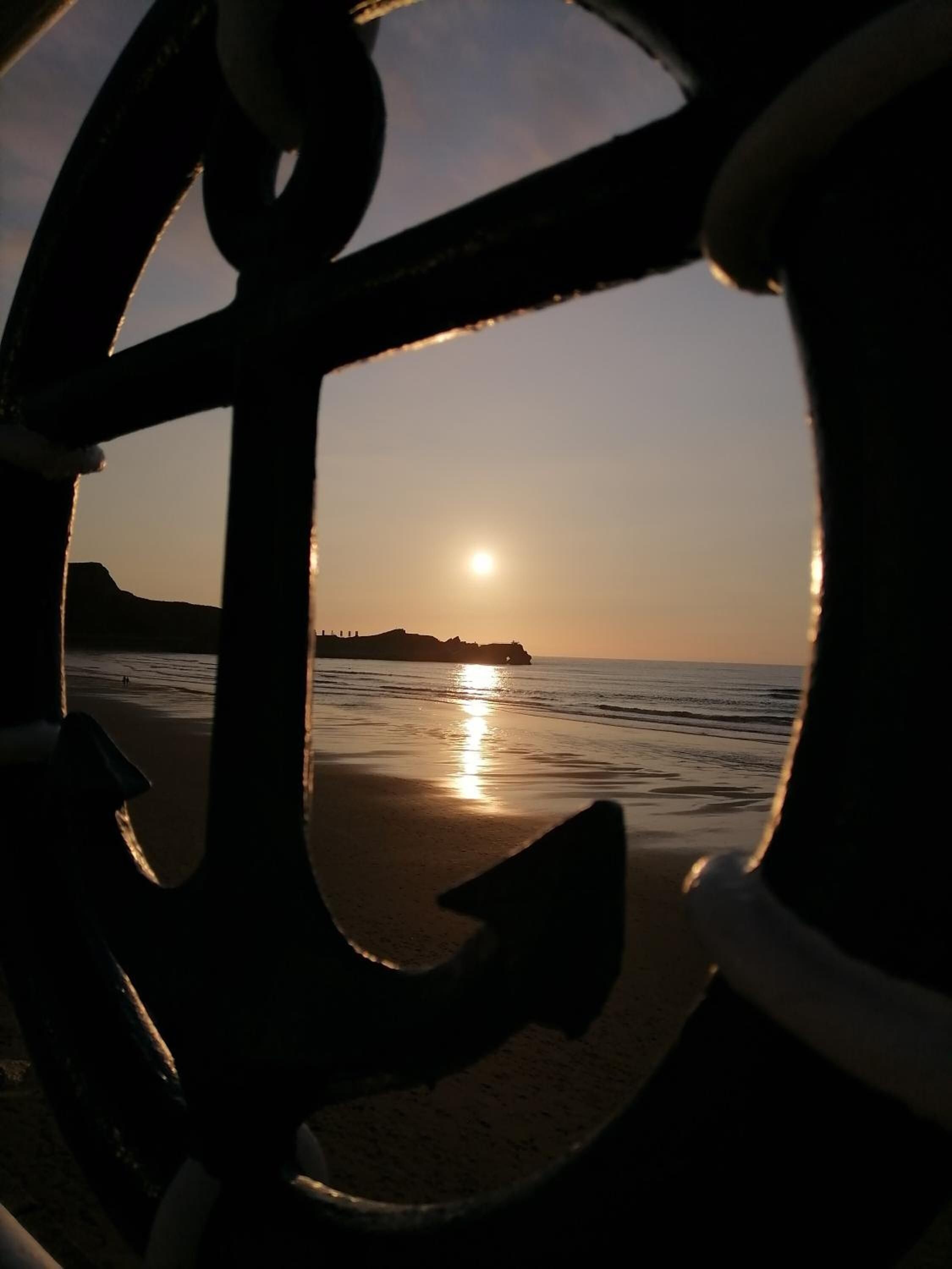 A sunset view through a metal anchor, with the sun setting over a beach and distant landforms.