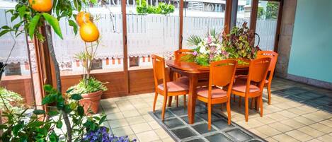 A covered patio area with a wooden table and chairs, surrounded by potted plants and a view of a building in the background.