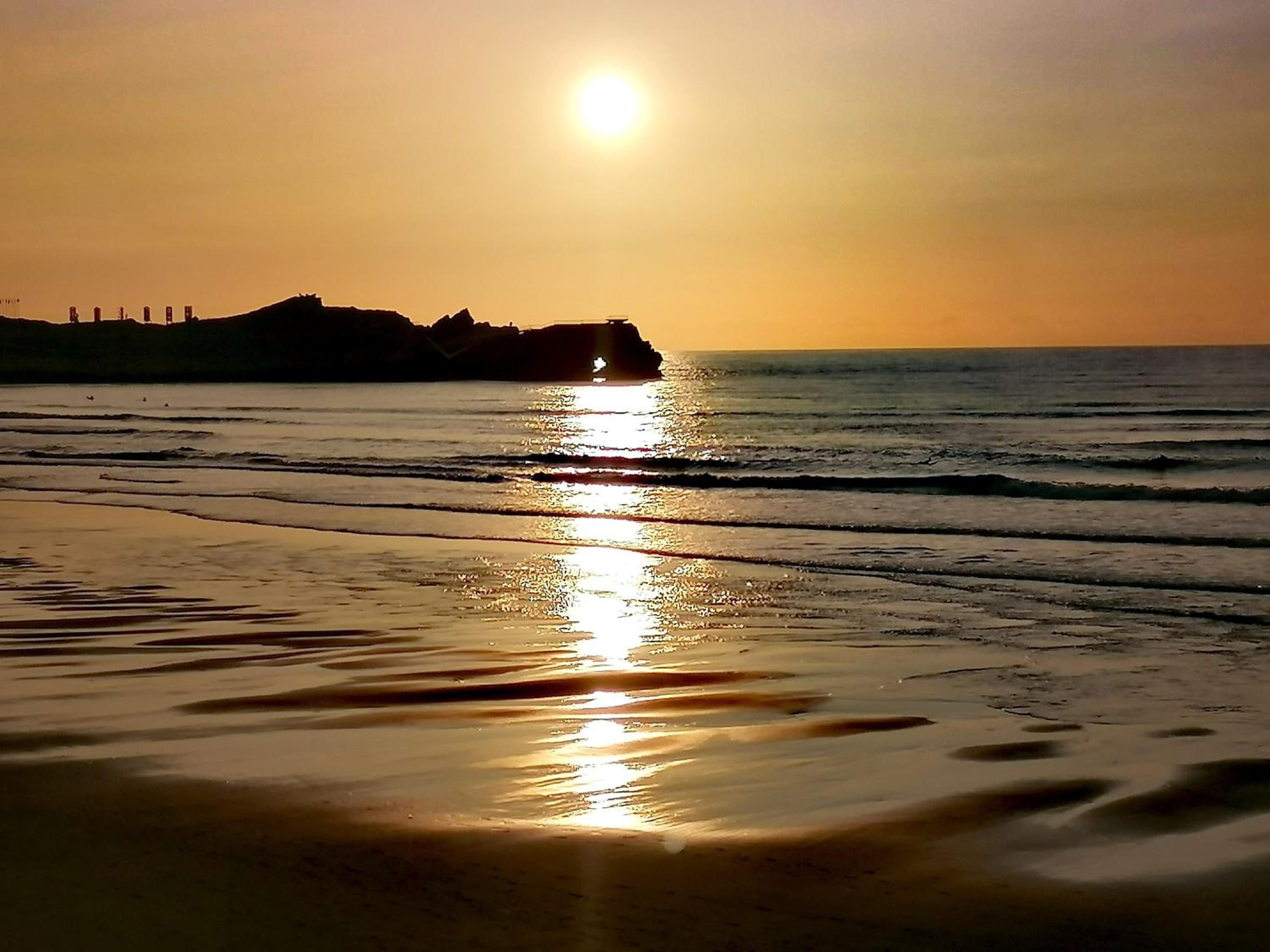 Ein Strand mit Sonnenuntergang, Wellen und einer fernen Felsformation.