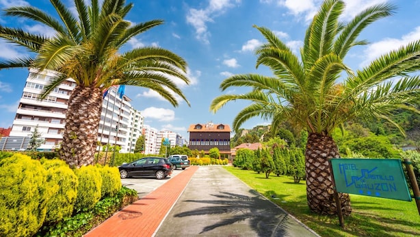 A parking area with palm trees, a pathway, and a building with a sign. - Hotel Castillo De Gauzón (Castrillon)