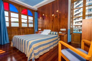A hotel room with a wooden paneled wall, a bed with a striped bedspread, a wooden chair, and a view of a building through a window with a blue curtain. - Hotel Castillo De Gauzón (Castrillon)