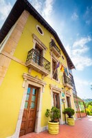 A yellow building with a traditional architectural style, featuring a balcony with ornate metal railings, a wooden door, and potted plants on the ground level.