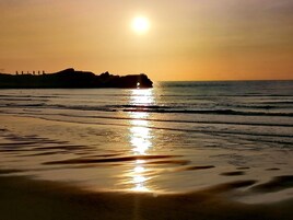 Praia com pôr do sol, ondas e formação rochosa ao longe.