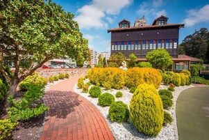 Um jardim ajardinado com um caminho curvo de tijolos, arbustos bem aparados e um edifício com design moderno ao fundo.