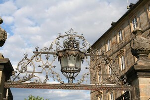 Una ornamentada puerta de hierro forjado con un farol, sobre un fondo de arquitectura clásica.