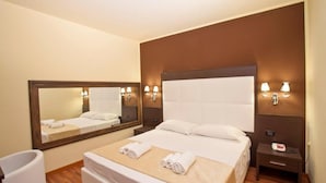 A hotel room with a large bed, a bedside table, a nightstand, a mirror, and a wall-mounted lamp. - Momi's Hotel (Cavarzere)
