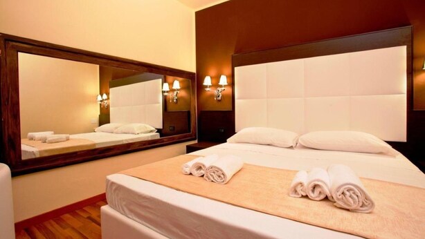 A hotel room with a large bed, a wooden headboard, a mirror, and wall-mounted lamps. - Momi's Hotel (Cavarzere)