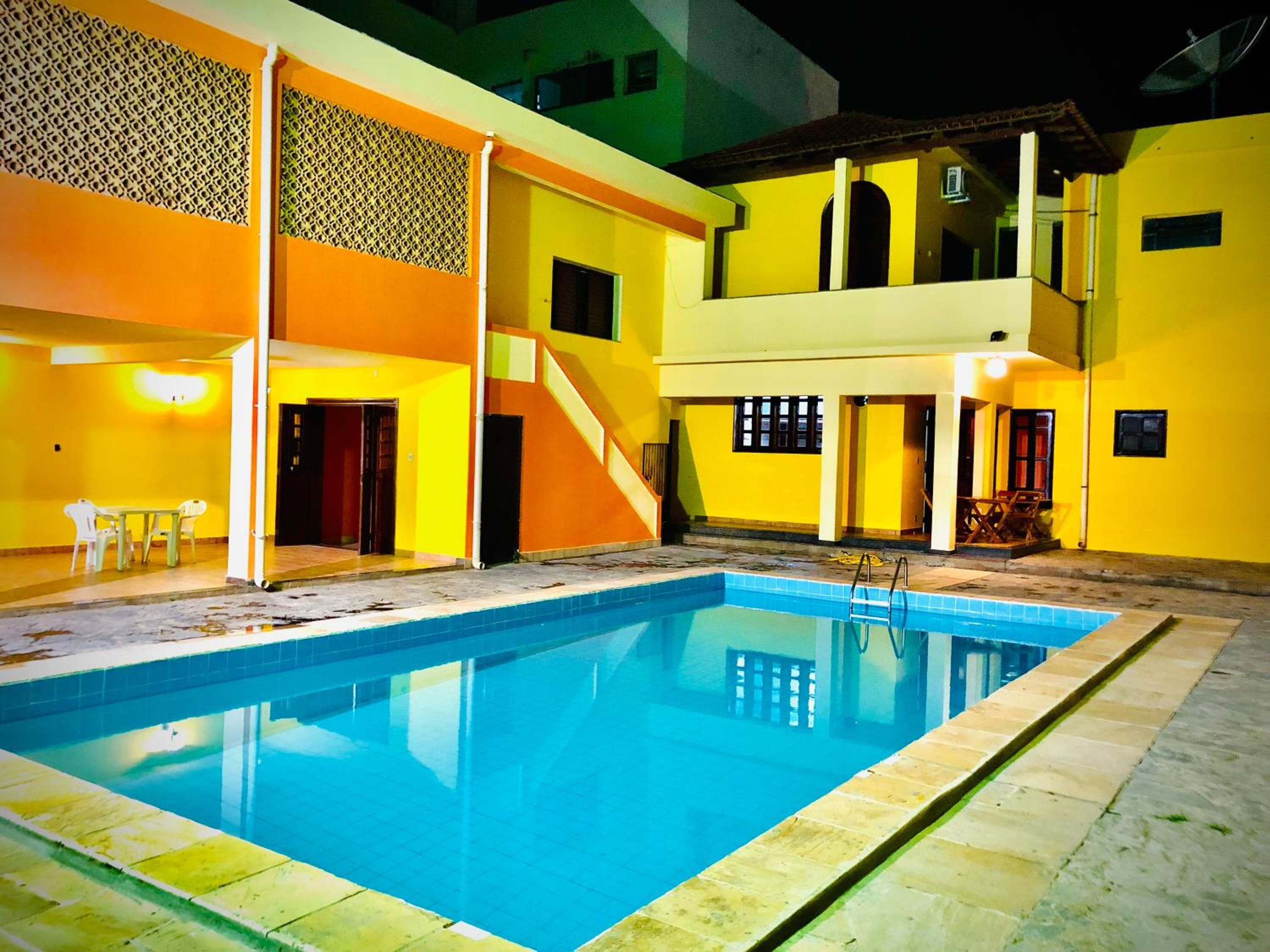 Una piscina con superficie de agua azul cristalina, rodeada por un edificio de paredes amarillas y una zona cubierta con mesa y sillas.
