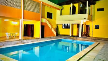 Piscina com superfície de água azul translúcida, rodeada por um edifício de paredes amarelas e uma área coberta com mesa e cadeiras.
