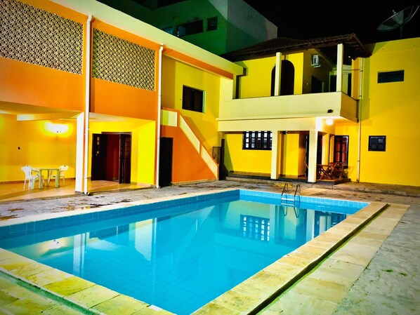 Una piscina con superficie de agua azul cristalina, rodeada por un edificio de paredes amarillas y una zona cubierta con mesa y sillas.