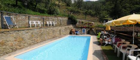A swimming pool with a stone wall and a patio area with chairs and umbrellas.