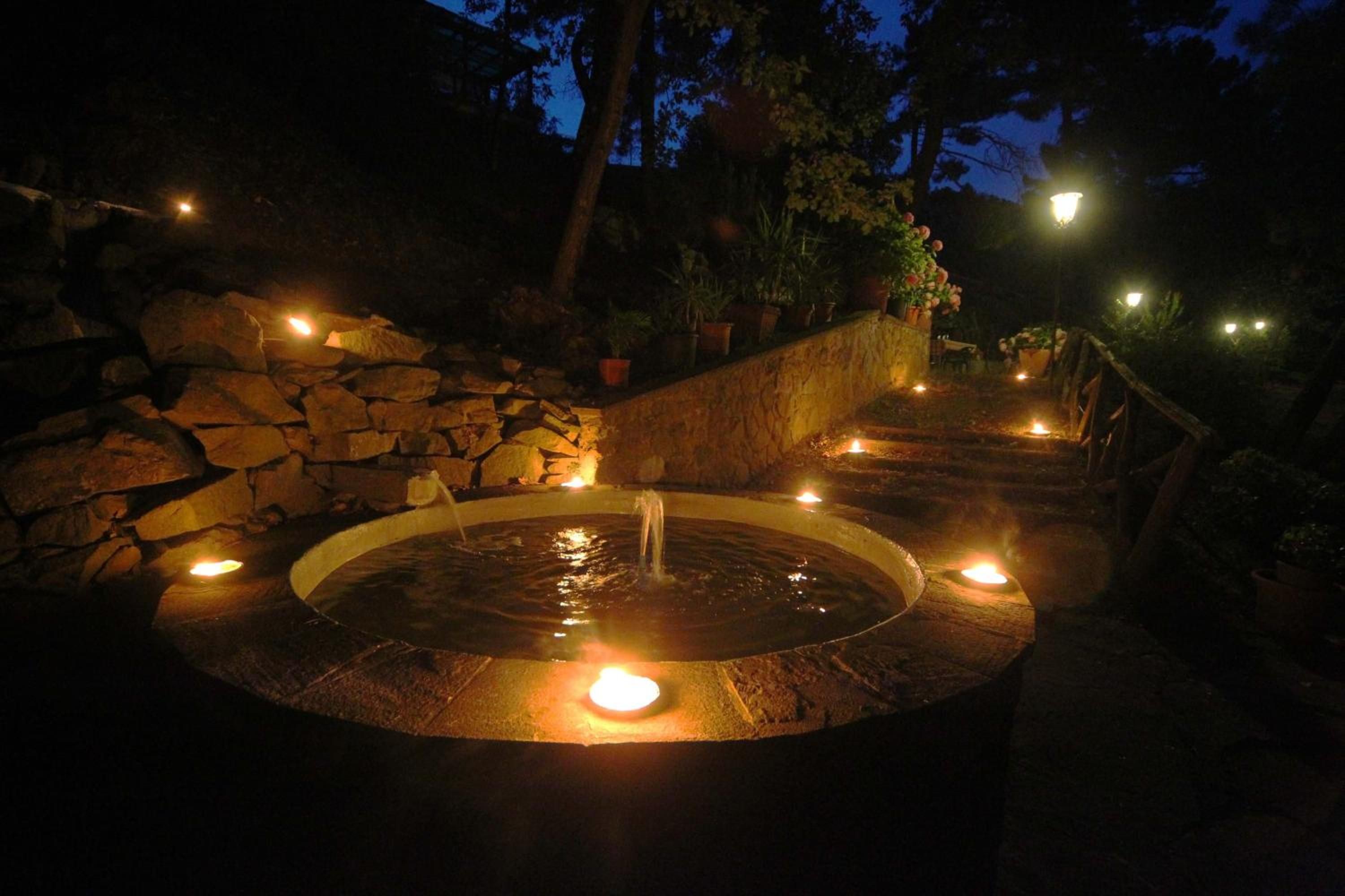 A circular hot spring with a stone wall and potted plants around it, illuminated by multiple lights.