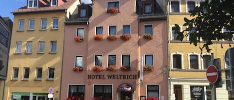 Un hôtel appelé « HOTEL WELTRICH », de style architectural européen traditionnel, se distingue par sa façade ornée de nombreuses fenêtres et de jardinières fleuries.