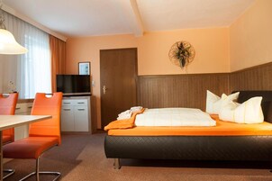 Quarto de hotel com uma cama, uma escrivaninha, uma cadeira, uma televisão e uma porta.
