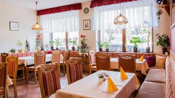 Ein Restaurant mit Holztischen und -stühlen, großen Fenstern mit Vorhängen und Topfpflanzen.