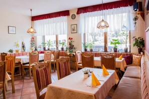 A restaurant with wooden chairs and tables, large windows with curtains, and potted plants.