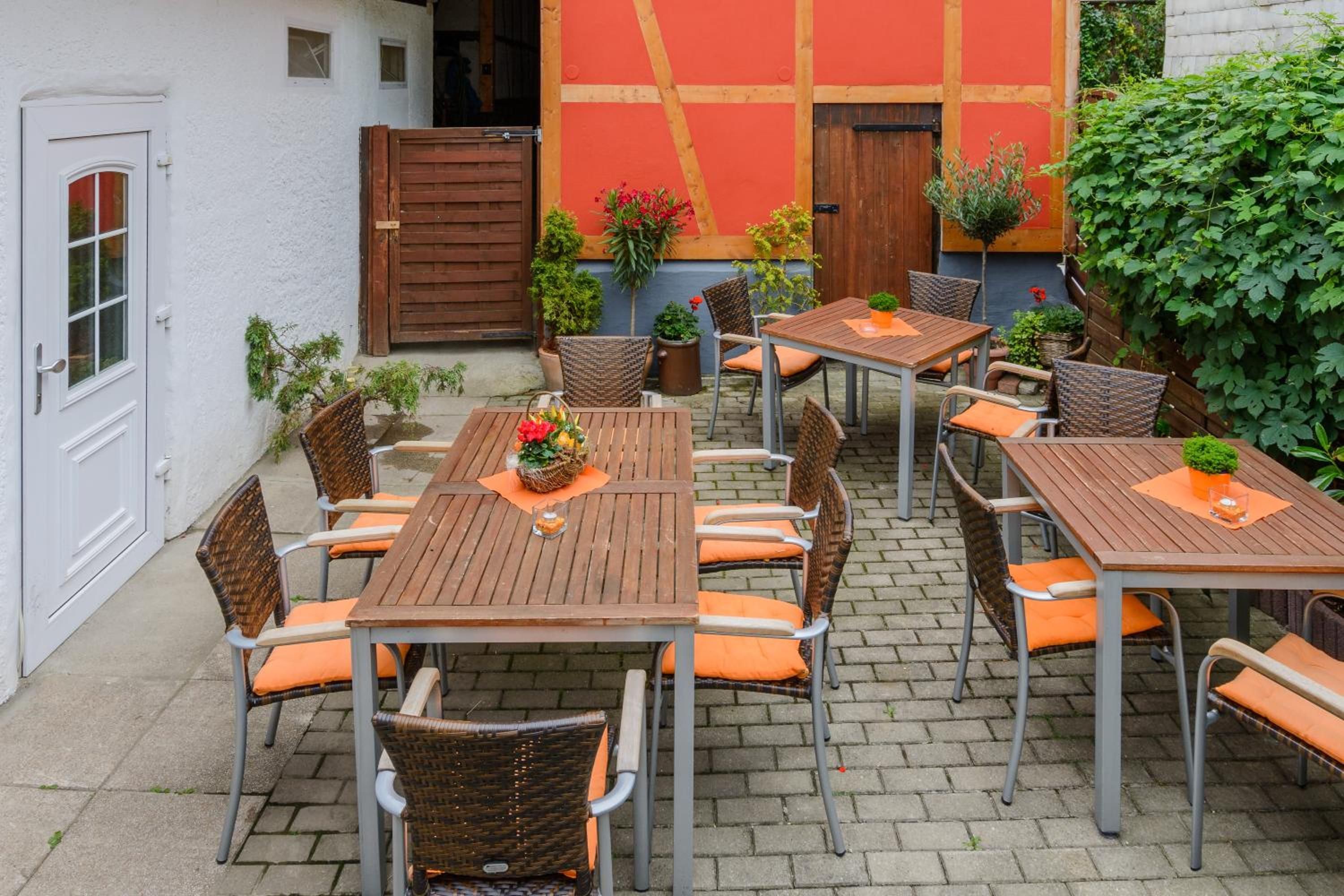An outdoor patio with multiple wooden tables and wicker chairs, surrounded by a white building with a red door and a wooden gate.