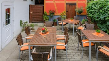 Eine Außenpatio mit mehreren Holztischen und Korbstühlen, umgeben von einem weißen Gebäude mit roter Tür und hölzernem Tor.