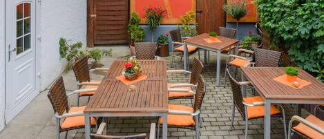 An outdoor patio with multiple wooden tables and wicker chairs, surrounded by a white building with a red door and a wooden gate.