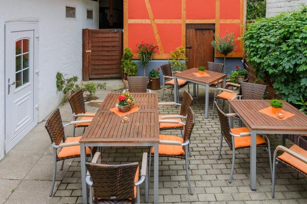An outdoor patio with multiple wooden tables and wicker chairs, surrounded by a white building with a red door and a wooden gate.