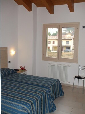 A bedroom with a bed, a bedside table with a lamp, a radiator, and a window with a view of buildings.