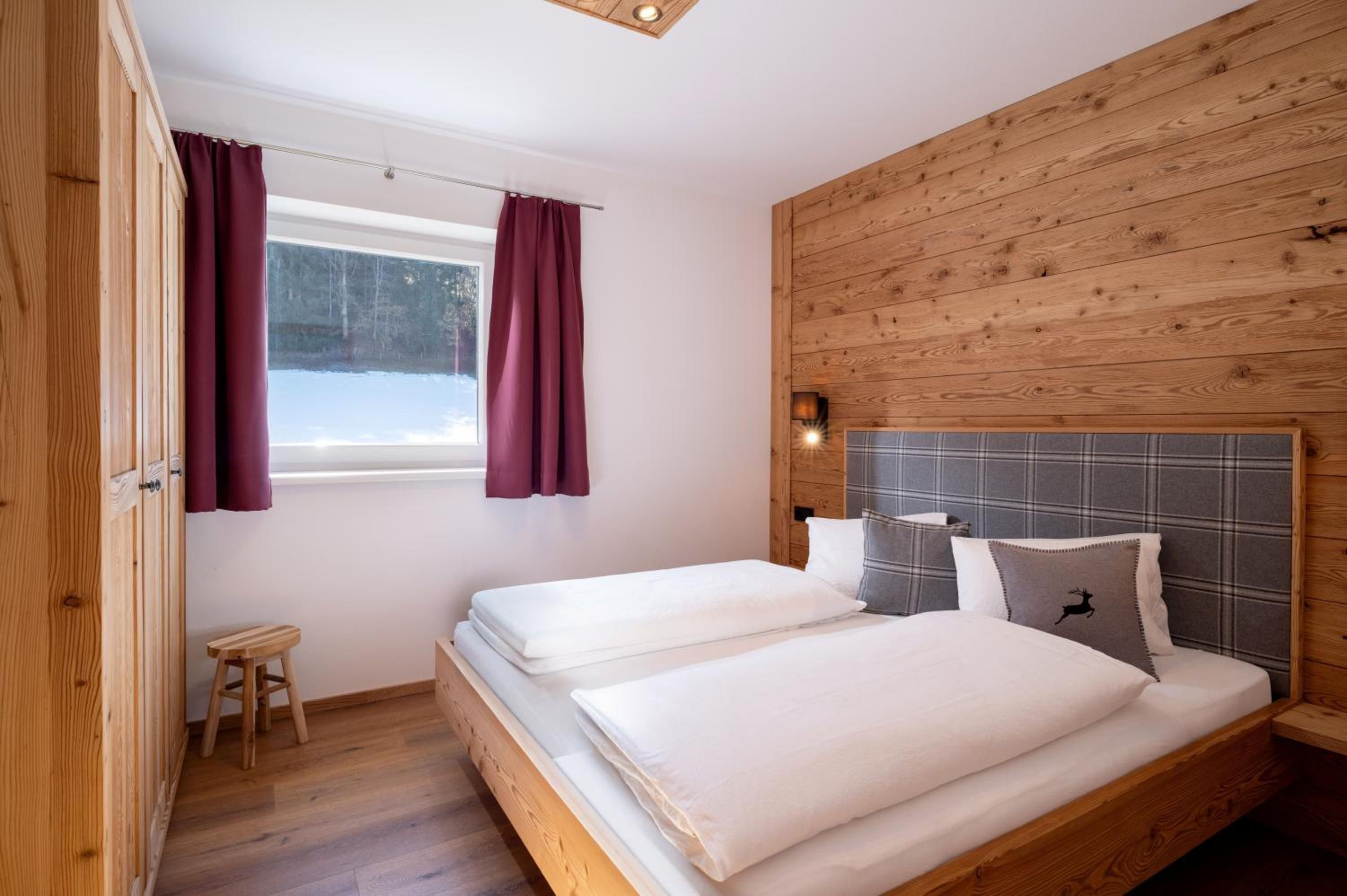 Quarto com duas camas, paredes de madeira, um banquinho pequeno e uma janela com cortinas.