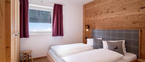 Quarto com duas camas, paredes de madeira, um banquinho pequeno e uma janela com cortinas.