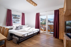 Ein Schlafzimmer mit Bett, Fernseher, Holzboden und einem Balkon mit Blick auf Häuser.