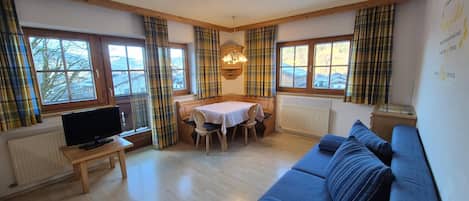 A cozy room with a wooden floor, a blue sofa, a dining table with chairs, a television on a wooden table, and large windows with plaid curtains.