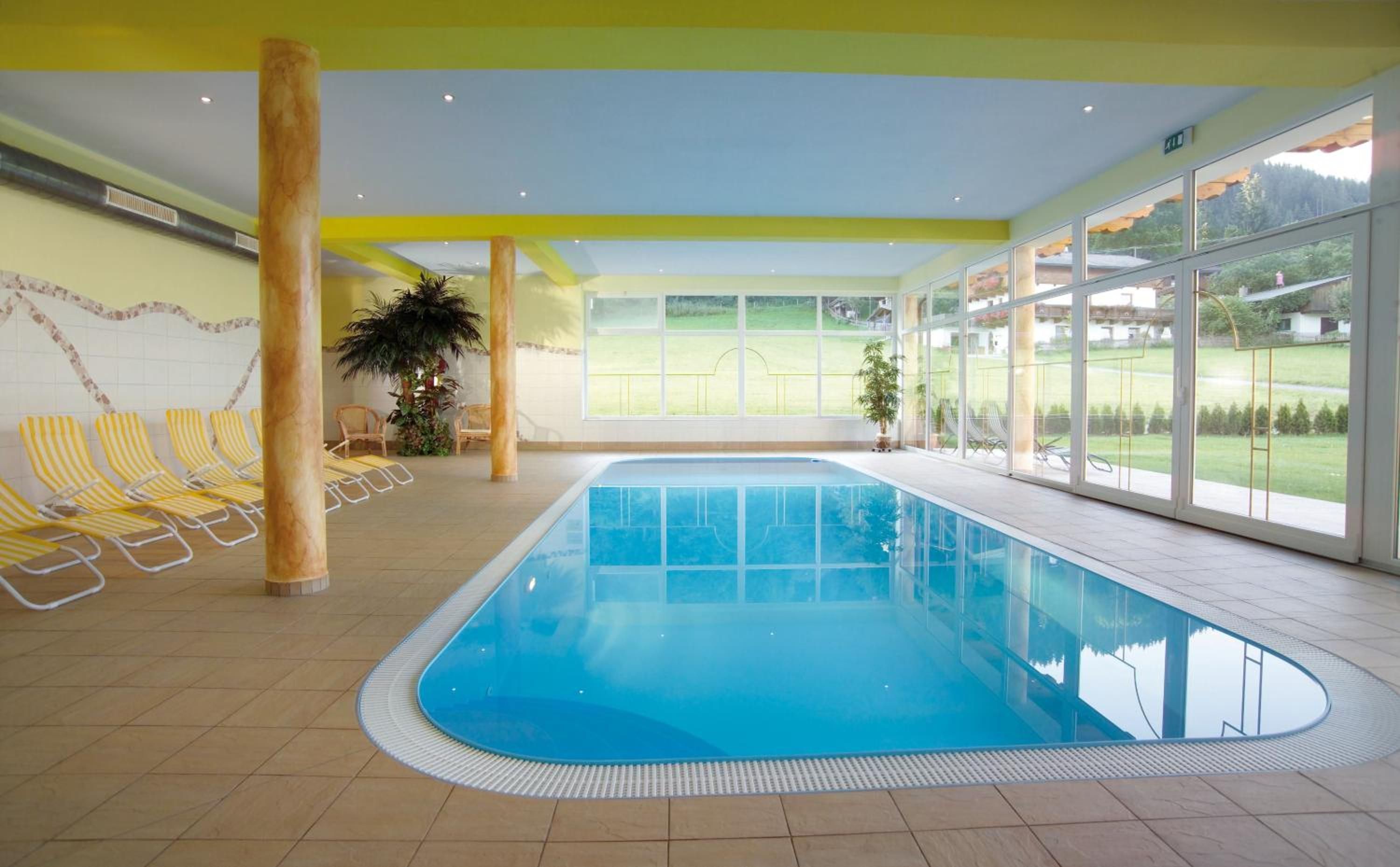 An indoor swimming pool with clear glass walls, yellow-striped lounge chairs, and a view of a green landscape.