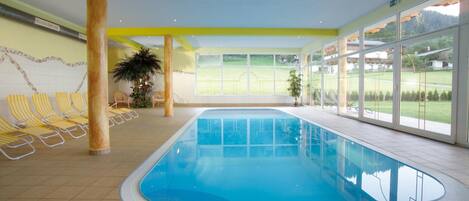 An indoor swimming pool with clear glass walls, yellow-striped lounge chairs, and a view of a green landscape.