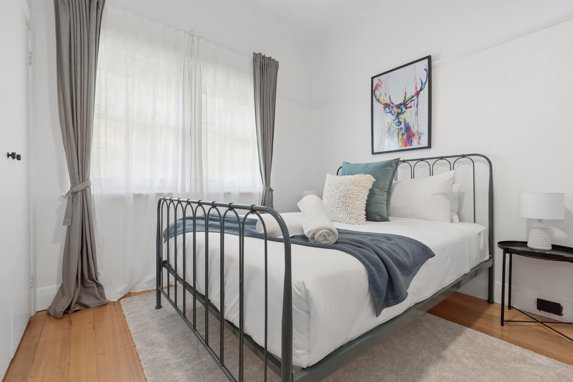 A bedroom with a metal bed frame, white bedding, a blue throw blanket, and a single bedside table with a lamp.