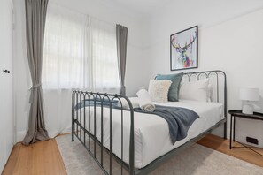 Un dormitorio con estructura de cama metálica, ropa de cama blanca, una manta azul y una mesita de noche con una lámpara.