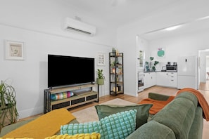 A modern living room with a flat-screen TV, a bookshelf, a sofa with colorful cushions, and a kitchen in the background.