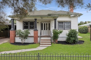 A single-story house with a front porch, a chimney, and a small front yard with two shrubs.