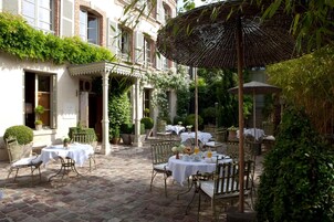 An outdoor patio with tables and chairs, surrounded by greenery and a building with brick walls and windows.