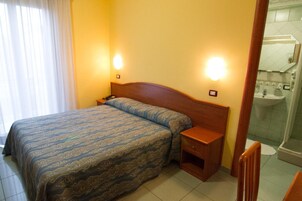 Habitación de hotel con cama, mesitas de noche, un tocador, un espejo y baño con lavamanos y espejo.