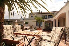 En udendørs terrasse med et træbord og stole, omgivet af plantekrukker og en bygning med mange vinduer.