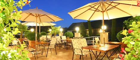 An outdoor patio area with tables and chairs, large umbrellas, and potted plants.
