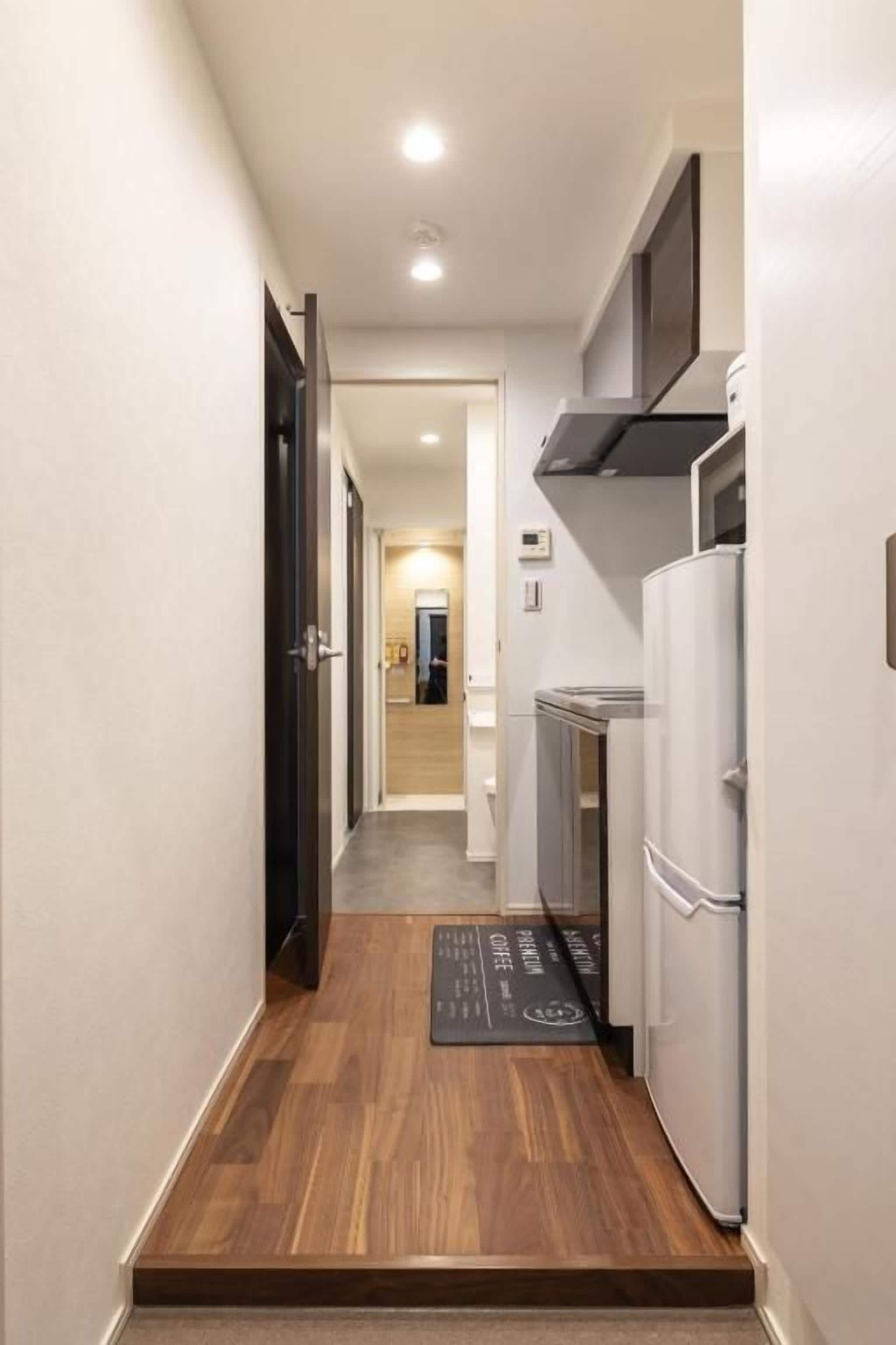 A narrow hallway with a wooden floor leading to a kitchen area with a refrigerator and cabinets.