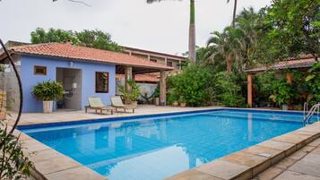 Piscina com borda em azulejo, rodeada por um terraço com uma espreguiçadeira e um pequeno edifício com telhado em azulejo.