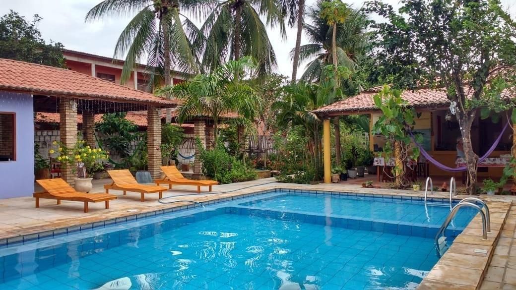 Piscina com espreguiçadeiras e área coberta, rodeada por palmeiras e um edifício com telhado em azulejos.