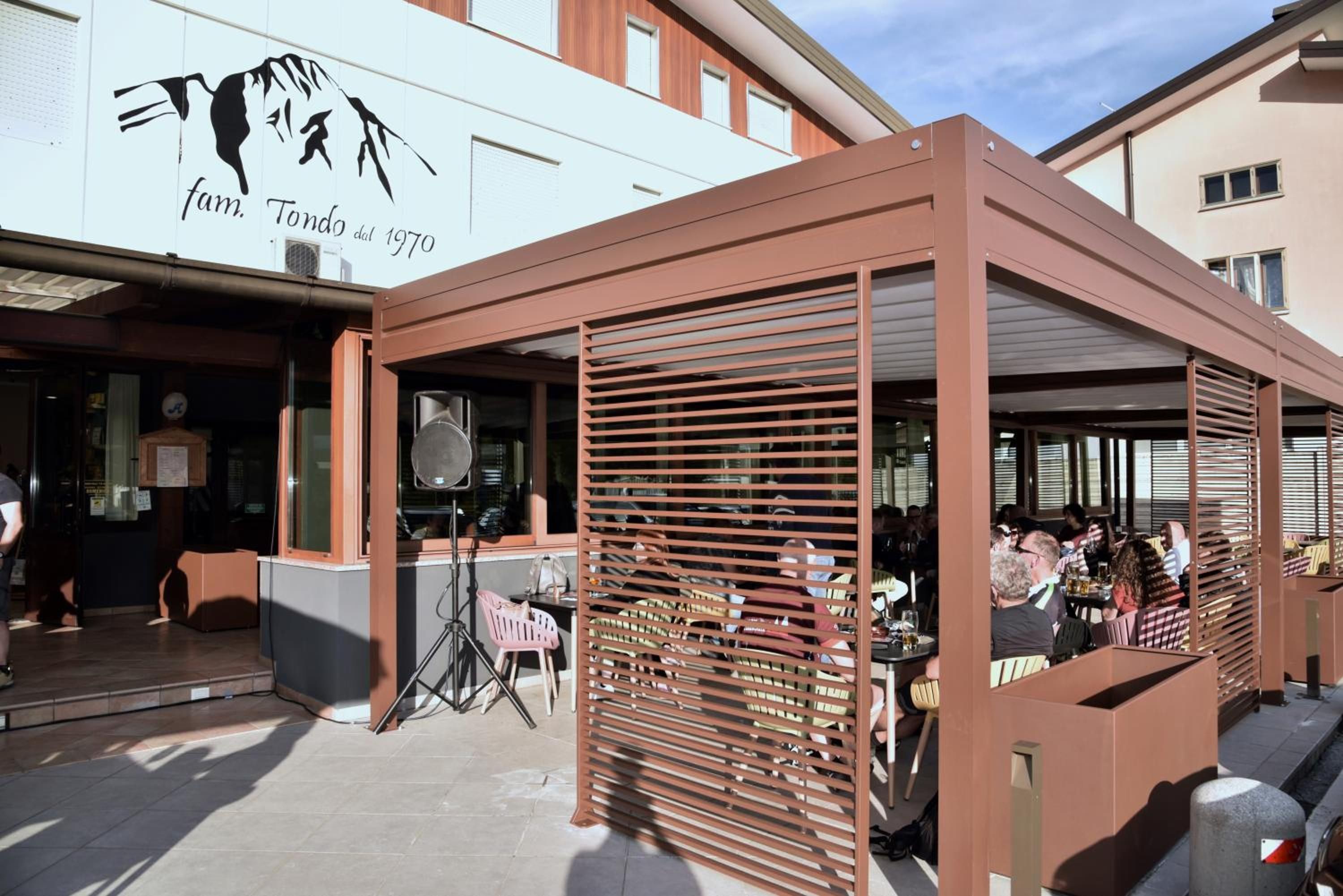 Un espace extérieur aménagé avec des tables et des chaises, une partie couverte avec un toit à lattes, et un bâtiment sur lequel on peut lire l’inscription suivante : « Ferme Tondo, depuis 1970 ».